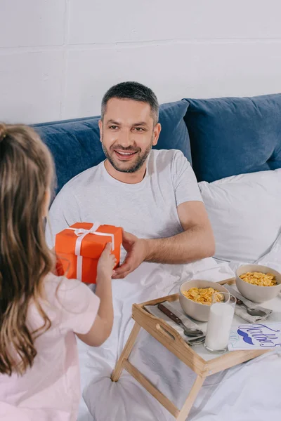 Back view of cute child presenting fathers day gift box to smiling father having breakfast in bed — Stock Photo