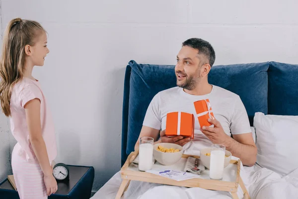 Happy man holding fathers day gift boxes while having breakfast in bed near adorable daughter — Stock Photo