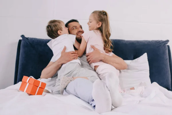 Adorable kids hugging happy dad on fathers day near gift box on bedding — Stock Photo