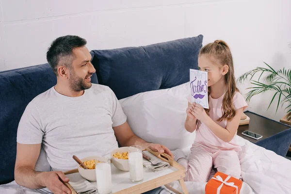 Cute daughter covering face with fathers day greeting card near smiling father holding tray with breakfast — Stock Photo