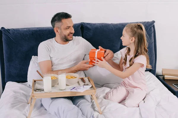 Lindo niño presentando padres día caja de regalo a papá desayunando en la cama - foto de stock