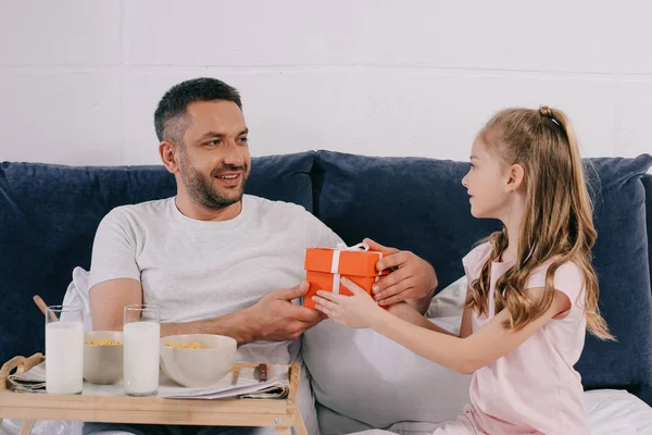 Adorable daughter presenting fathers day gift box to smiling dad having breakfast in bed — Stock Photo