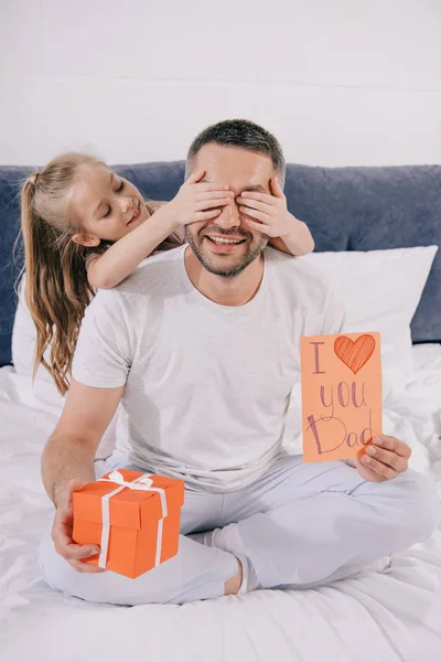 Smiling man holding gift box and fathers day greeting card while cheerful daughter covering his eyes with hands — Stock Photo