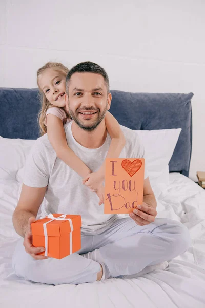 Bambino sorridente abbracciando felice papà in possesso di scatola regalo e papà giorno biglietto di auguri — Foto stock