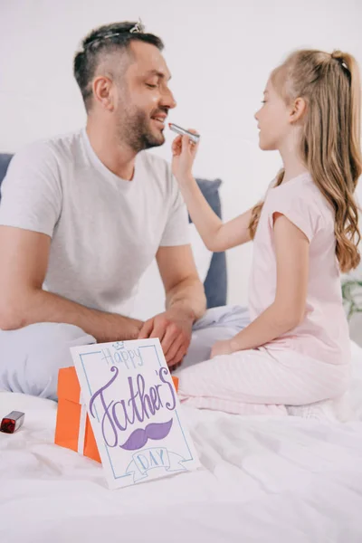 Lindo niño aplicando lápiz labial en los labios del padre cerca de la caja de regalo y la tarjeta de felicitación del día del padre en la ropa de cama - foto de stock