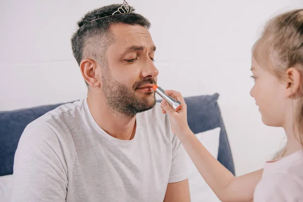 Happy man in diadem sitting with closed eyes while adorable daughter applying lipstick on his lips — Stock Photo