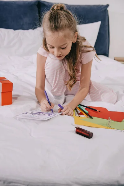 Lindo niño dibujo en los padres tarjeta de felicitación día mientras está sentado en la ropa de cama - foto de stock