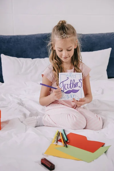 Adorable niño dibujo padres día tarjeta de felicitación mientras está sentado en la ropa de cama cerca de papel multicolor - foto de stock