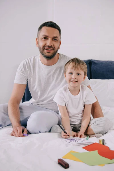 Feliz hombre sentado en la ropa de cama cerca de adorable hijo haciendo padres tarjeta de felicitación día - foto de stock