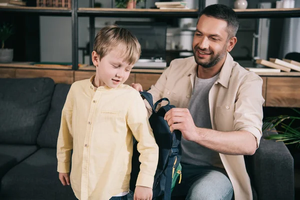 Smiling dad helping cute boy putting on backpack at home — Stock Photo