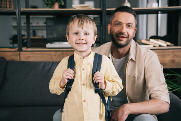 Niño sonriente con mochila en los hombros sonriendo a la cámara junto con papá feliz - foto de stock