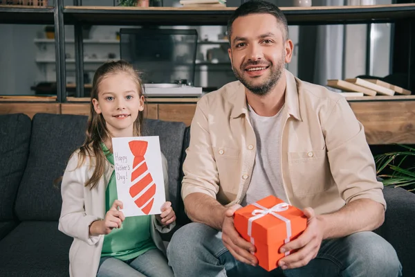 Heureux père tenant boîte-cadeau tout en étant assis près de fille adorable présentant la carte de voeux de jour des pères avec cravate tirée — Photo de stock