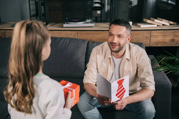 Selective focus of daughter presenting gift box to smiling dad holding fathers day greeting card with drawn tie — Stock Photo