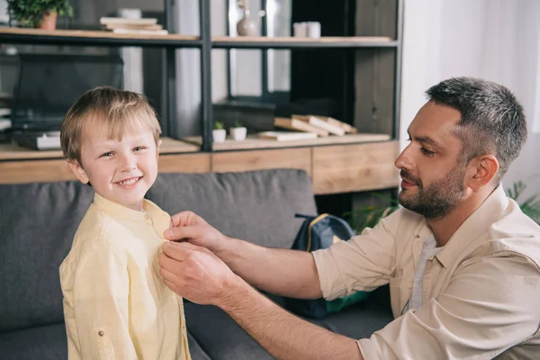 Feliz hombre ayudando a vestir sonriente hijo en casa - foto de stock