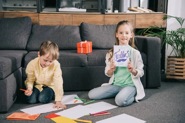 Enfant mignon montrant la carte de voeux de jour de pères tout en étant assis sur le sol près du frère dessin sur des feuilles de papier blanc — Photo de stock