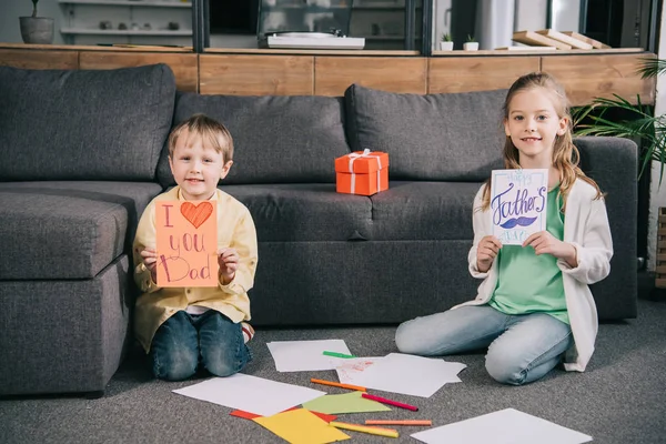 Adorables niños mostrando tarjetas de felicitación de día de padres hechos a mano mientras están sentados en el suelo cerca del sofá - foto de stock