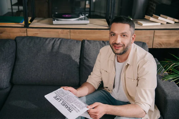 Schöner bärtiger Mann, der in die Kamera lächelt, während er auf dem Sofa sitzt und eine Reisezeitung in der Hand hält — Stockfoto