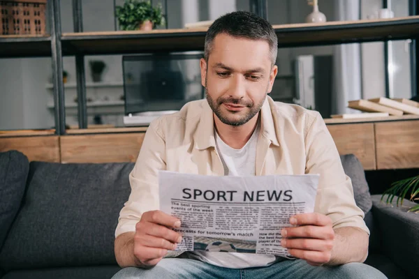 Bonito homem ler esporte notícias jornal enquanto sentado no sofá em casa — Fotografia de Stock