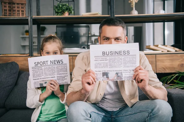 Alegre hija y padre cubriendo caras con periódicos de negocios y noticias deportivas - foto de stock