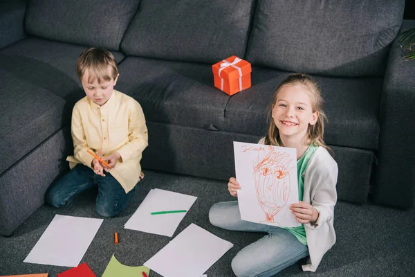 Alegre chica mostrando dibujo y sonriendo a la cámara mientras está sentado en el suelo cerca de hermano - foto de stock