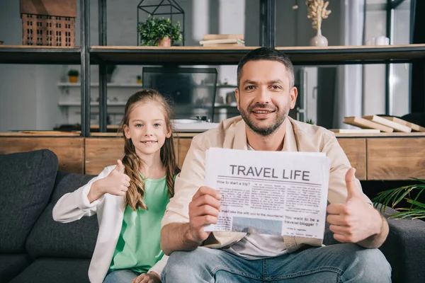 Smiling father and daughter showing thumbs up while sitting on sofa with travel life newspaper — Stock Photo