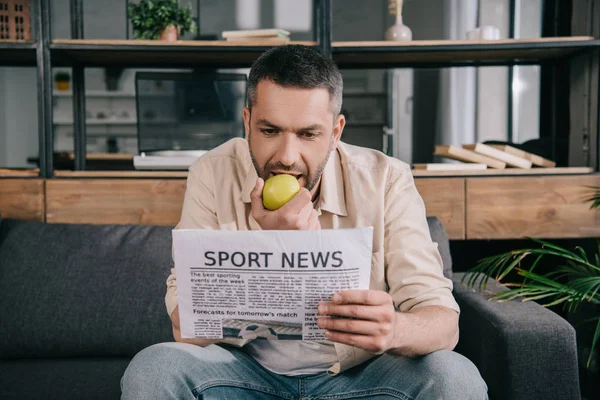 Handsome man eating fresh apple while reading sport news newspaper at home — Stock Photo