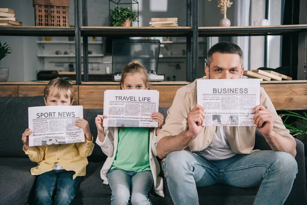 Alegre padre e hijos sosteniendo periódicos mientras están sentados en el sofá en casa - foto de stock