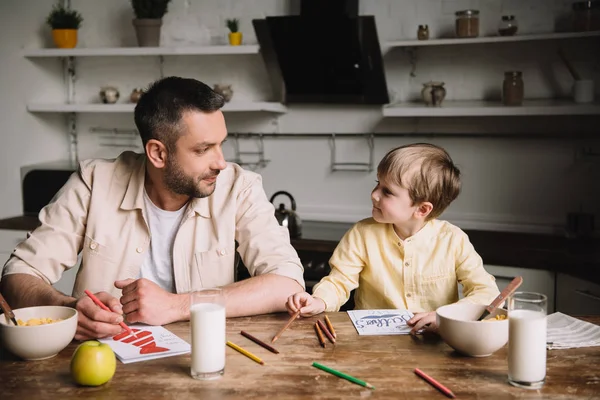 Pai e filho desenho enquanto sentado na mesa da cozinha com café da manhã servido — Fotografia de Stock