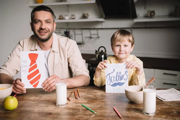 Feliz padre e hijo mostrando tarjetas de felicitación día padres mientras está sentado en la mesa de la cocina con el desayuno servido - foto de stock