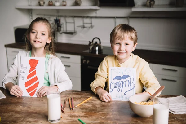 Sonriente hermano y hermana mostrando a los padres tarjetas de felicitación día mientras está sentado en la mesa de la cocina con el desayuno servido - foto de stock