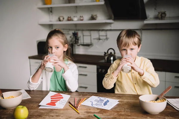 Entzückende Kinder, die Milch trinken, während sie am Küchentisch neben handgemachten Vatertagsgrüßkarten sitzen — Stockfoto