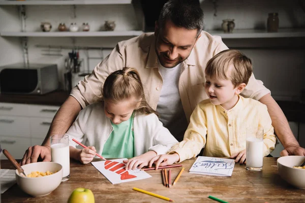 Glücklicher Papa, der neben entzückenden Kindern steht und Vatertagsgrüßkarten zeichnet, während er am Küchentisch mit serviertem Frühstück sitzt — Stockfoto