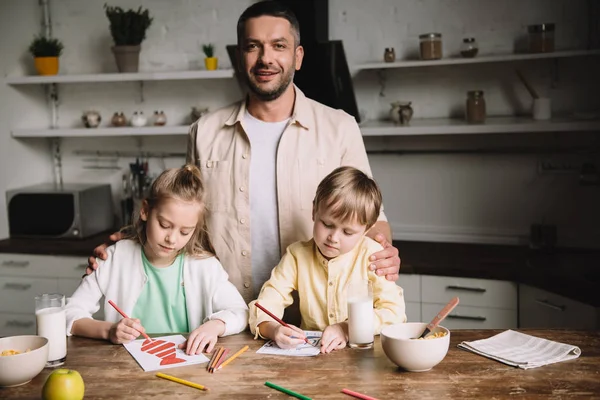 Happy father standing near adorable kids drawing fathers day greeting cards at kitchen table — Stock Photo