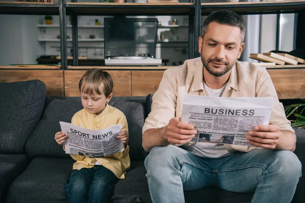 Concentrato padre e figlio leggere giornali mentre seduto sul divano a casa — Foto stock
