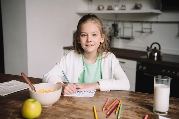 Niedliches Kind lächelt in die Kamera, während es am Küchentisch sitzt und eine Grußkarte zum Vatertag zeichnet — Stockfoto