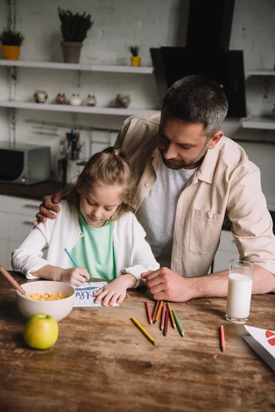 Happy father hugging adorable daughter sitting at kitchen table with served breakfast and drawing fathers day greeting card — Stock Photo