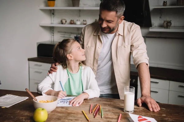 Glücklicher Vater umarmt entzückende Tochter sitzt am Holztisch mit serviertem Frühstück und zeichnet Vatertags-Grußkarte — Stockfoto