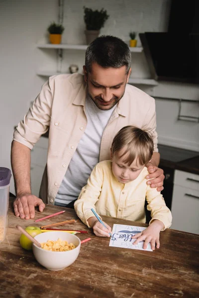 Glücklicher Vater umarmt entzückenden Sohn sitzt am Küchentisch und zeichnet Vatertags-Grußkarte — Stockfoto