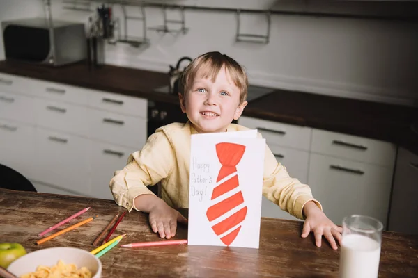 Carino ragazzo mostrando padri giorno biglietto di auguri con cravatta disegnata mentre seduto al tavolo della cucina e sorridente alla fotocamera — Foto stock