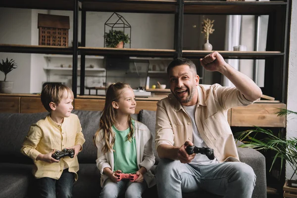 KYIV, UCRANIA - 10 de mayo de 2019: Familia feliz jugando videojuegos con joysticks mientras padre muestra el gesto ganador . - foto de stock