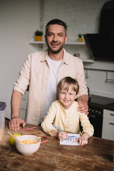 Feliz padre abrazando adorable hijo sentado en la mesa de la cocina y dibujando los padres tarjeta de felicitación día - foto de stock