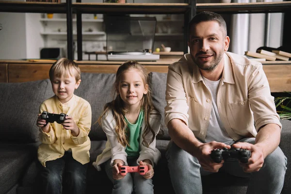 KYIV, UCRAINA - 10 MAGGIO 2019: Buon padre e adorabili bambini che giocano a videogiochi con joystick a casa . — Foto stock