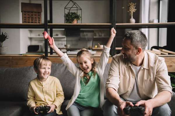 KYIV, UCRAINA - 10 MAGGIO 2019: Bambino eccitato che festeggia il trionfo seduto vicino a padre e fratello con i joystick in mano . — Foto stock