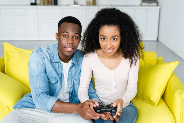 KYIV, UKRAINE - 13 MAI 2019 : vue en grand angle du couple afro-américain assis sur le canapé et tenant une manette de jeu dans les mains — Photo de stock