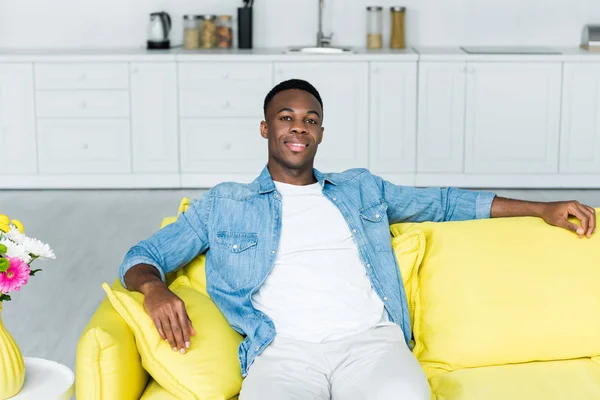 Africano americano uomo sorridente, seduto sul divano e guardando la fotocamera — Foto stock