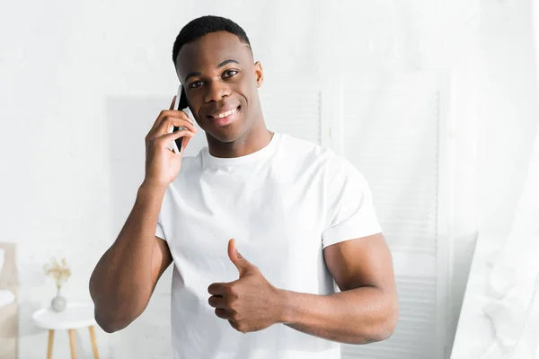 Hombre afroamericano feliz hablando en smartphone y mostrando el pulgar hacia arriba - foto de stock