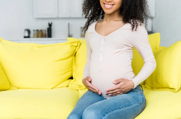 Cropped view of pregnant african american woman sitting on couch at home — Stock Photo