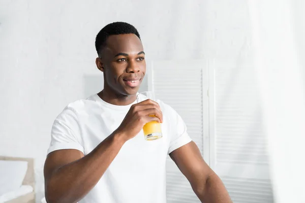 Hombre afroamericano feliz mirando hacia otro lado y bebiendo jugo de naranja - foto de stock