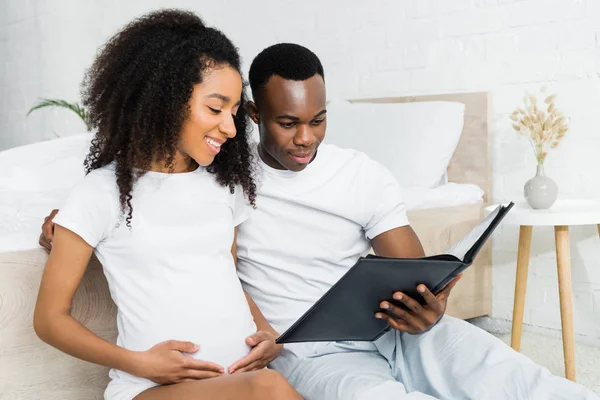 Mujer afroamericana embarazada sentada cerca de novio, mirando álbum de fotos - foto de stock