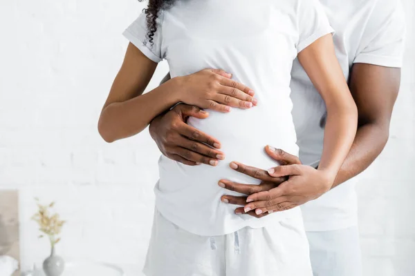 Recortado vista de africano americano hombre abrazando embarazada esposa en habitación - foto de stock